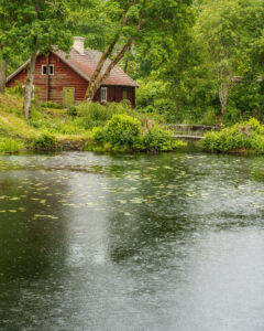 Röd faluröd stuga vid en stilla damm med näckrosblad, omgiven av frodig grönska vid Håfla hammarsmedja i Östergötland