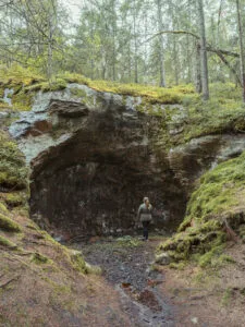 Kvinna står vid en stor jättegryta som liknar en grotta bland mossbeklädd skog vid Jättegrytorna i Steneby, Dalsland.