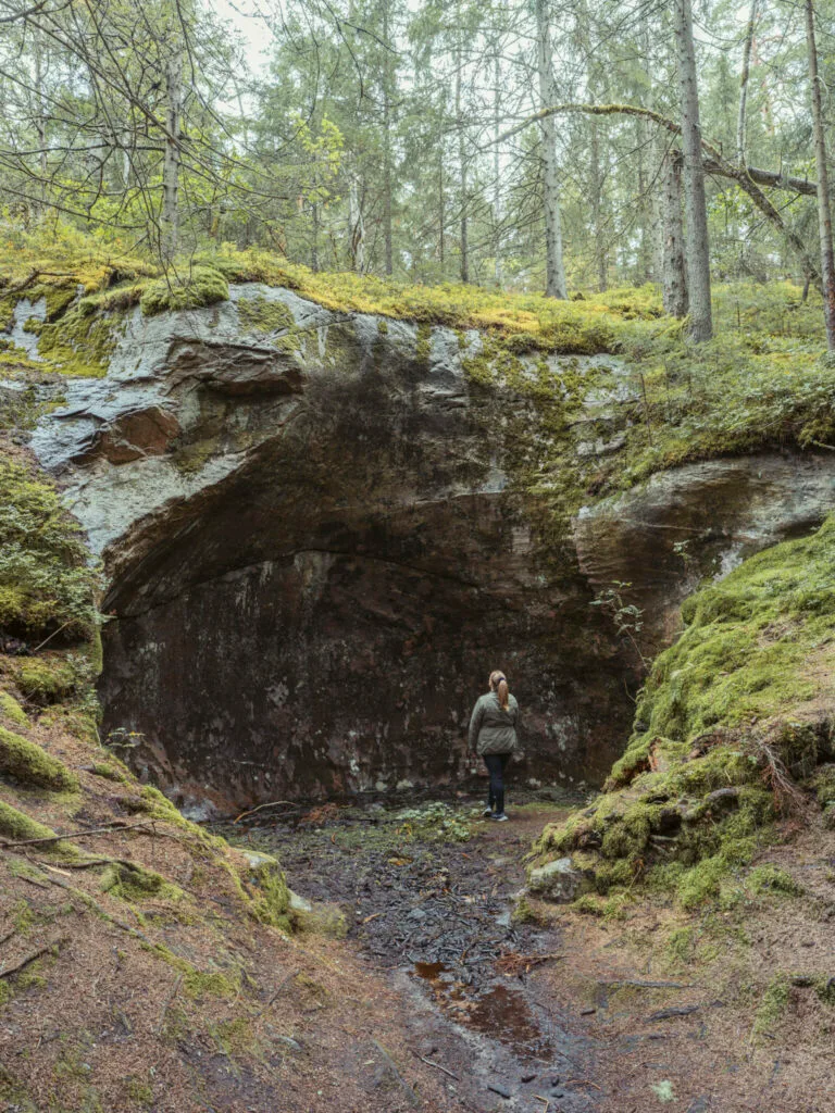 Kvinna står vid en stor jättegryta som liknar en grotta bland mossbeklädd skog vid Jättegrytorna i Steneby, Dalsland.