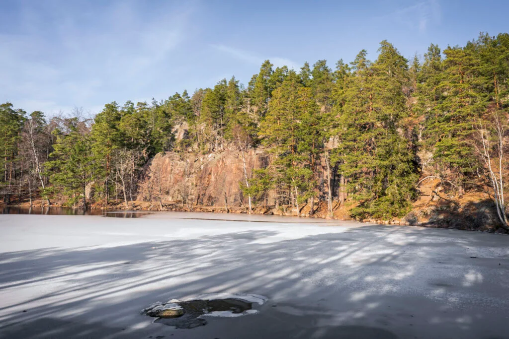 Isbelagd sjö med speglande skuggor framför branta klippor och tallskog en solig vinterdag vid Ageljön.
