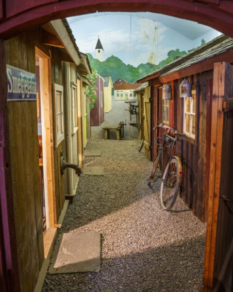 Museiutställning på Melleruds museum med rekonstruerad gränd vid Smedjegatan, gammalt trähus och cykel mot väggen.