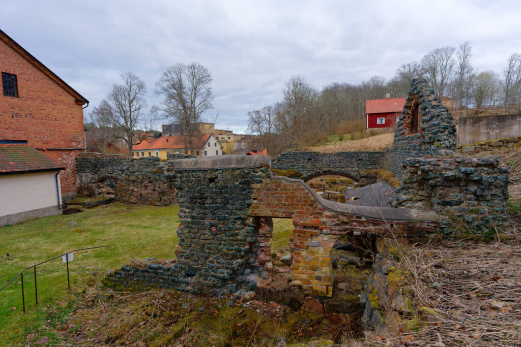 Ruiner av den gamla smedjan och masugnsgrunden i Edsbruk, med stenmurar, tegelbyggnad och herrgårdsmiljö i bakgrunden.