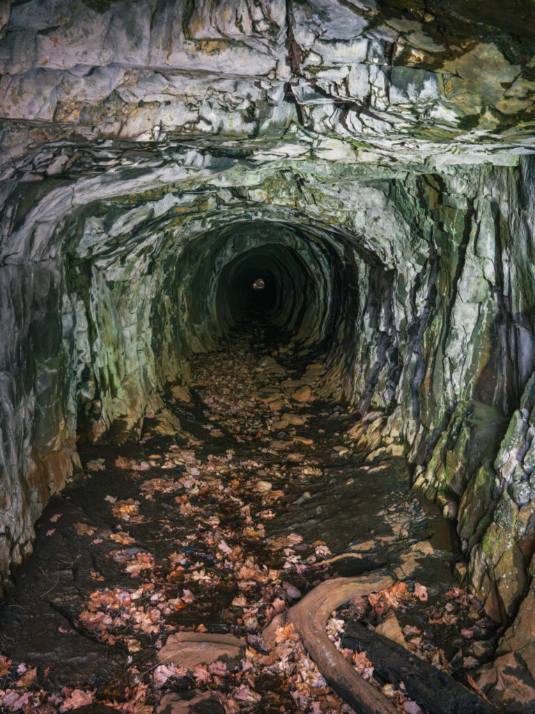 Mörk stollgång med råa bergväggar i Stenebo storgruva, med löv på marken och ett svagt ljus synligt längst in i tunneln